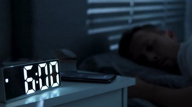 Man waking up, turning off alarm clock on digital clock and smartphone. Sleepy man waking up at 6 o'clock morning