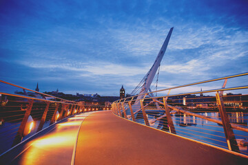 Derry, Ireland. Illuminated Peace bridge in Derry Londonderry, City of Culture, in Northern Ireland with city center at the background. Night cloudy sky with reflection in the river at the dusk