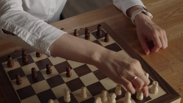 A business woman wearing a white shirt plays chess by herself in her home office. She contemplates her next move in the game.