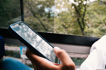 Hand of man using his phone on a bus with trees in the background and the reflection in the screen