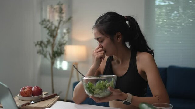 Athletic woman struggles to eat a salad meal while sitting at her dining table, showing clear signs of disgust and dissatisfaction