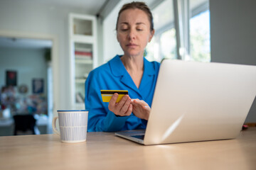 Serious woman types card info on laptop for online payments, risking financial issues. Credit concerns, spending overdrive, debt accumulation, insecure payment, shopping compulsion, online scams