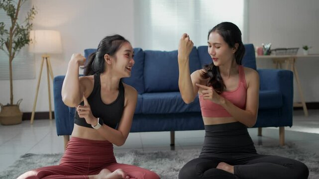 Two athletic women are comparing biceps progress while using a fitness app on a smartphone in a cozy living room