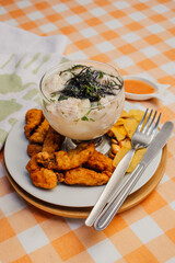 Leche de tigre presentado en copa, sobre un plato blanco acompañado de chifles, canchita y chicharrón de pescado, sobre mantel de cuadros naranja