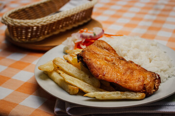 Pollo frito, acompañado de papas fritas y arroz blanco en plato blanco, sobre mesa a cuadritos anaranjado, con cubiertos a un lado