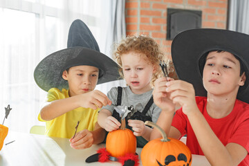 Three real siblings two brothers and sister making together jack o lantern at their home white kitchen, cutting real pumpkins and playing with paper black ghosts, lovely vibe of halloween