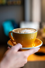 A close-up of a hand holding a yellow cup with a saucer, containing coffee with foam art on top. The background is blurred, emphasizing the cup and its contents. Perfect for themes of coffee