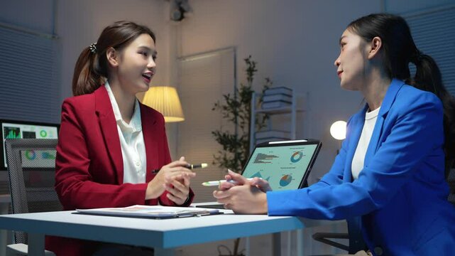 Two businesswomen working late in the office, discussing strategies and ideas over a digital tablet