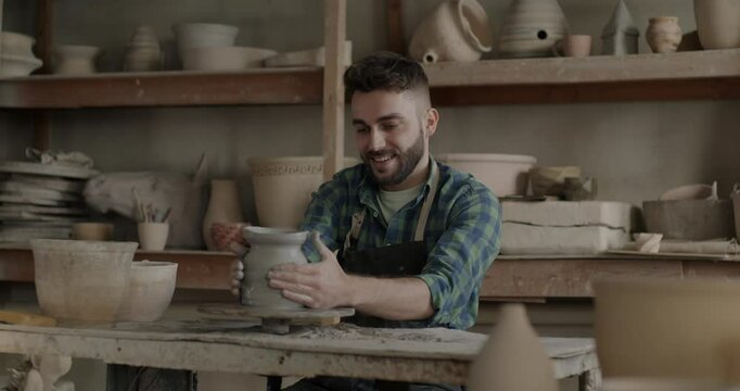 Slow motion of young man in dirty apron using spinning wheel enjoying pottery in workshop. Creative activity and positive emotion concept.