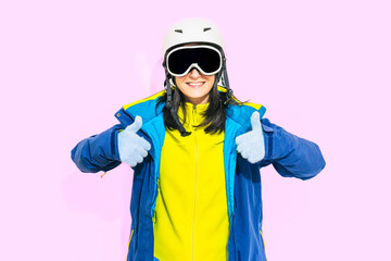 Close up portrait pretty young woman front view wear winter ski outfit goggles helmet gloves show thumbs up smiling isolated by pink studio background. Winter lifestyle, extreme sport preparations
