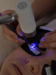 A woman undergoing eyelash extension procedure using an ultraviolet lamp.