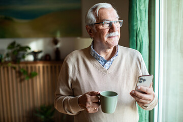 Senior man looking out window holding smartphone and coffee mug