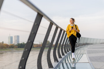 Cheerful woman strolls along a modern bridge, chatting on her phone while enjoying the serene...