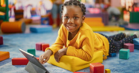 Happy African Girl Using Tablet Computer for Studying and Playing in Kindergarten. Beautiful Black Child Looking at Camera and Smiling. Happy Children Spending Time in a Modern Daycare Center