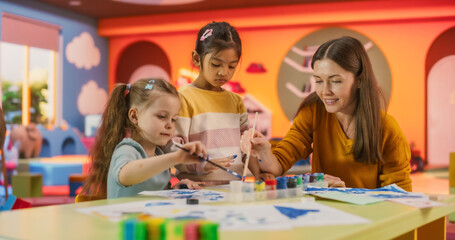 Fototapeta premium Portrait of Two Adorable Girls Using Watercolor to Create Colorful Paintings on Paper. Cheerful Female Teacher Spending Productive Time in Preschool, Teaching Kids to Paint in Art Class