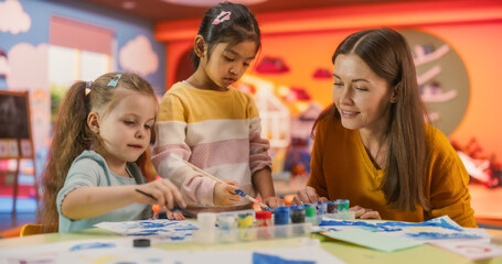 Fototapeta premium Portrait of Two Adorable Girls Using Watercolor to Create Colorful Paintings on Paper. Cheerful Female Teacher Spending Productive Time in Preschool, Teaching Kids to Paint in Art Class