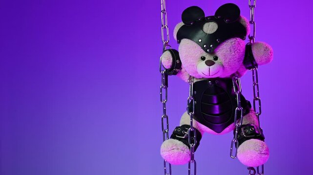 A teddy bear in a mask is suspended from chains in leather handcuffs and accessories for bdsm games and sex in neon light against a background of copy paste