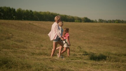 Side view of child walking with her two aunts through vast open field, one aunt holding rope while they enjoy outdoor stroll, surrounded by nature, lush green trees in background