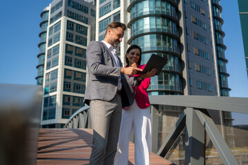 Smiling Latin Hispanic mature adult businesspeople using pc digital computer. Laughing caucasian colleagues, checking the company budget on their laptop in front of glass building exterior.	