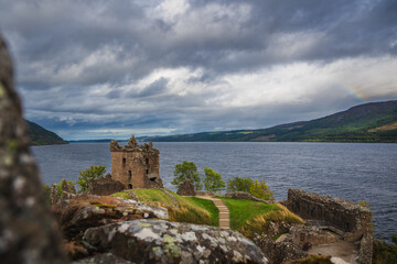 Ruins of Urquhart Castle in the Highlands of Scotland besides Loch Ness lake in background with dramatic sky and part of rainbow.