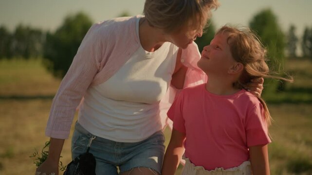 Mother and daughter walking happily in open field with mother smiling as she gently kisses daughter on cheek, mother holding bouquet of flowers in hand, enjoying sunny day