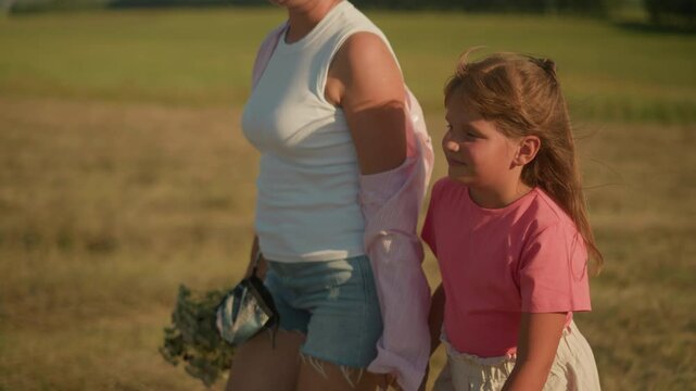 Side view of little girl and aunt walking hand in hand in sunny countryside, aunt holding bouquet of flowers, both smiling warmly, enjoying summer day in open field with blurred green landscape
