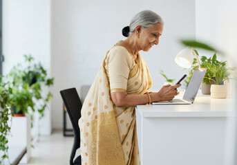 Elderly Indian woman using smartphone and laptop in minimalist home office with plants