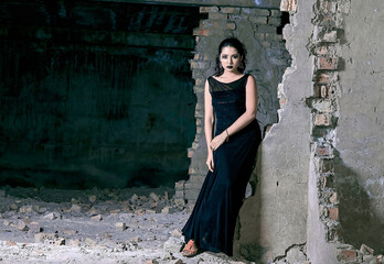 A beautiful young girl model stands in a long black dress amidst the rubble of a partially demolished building.