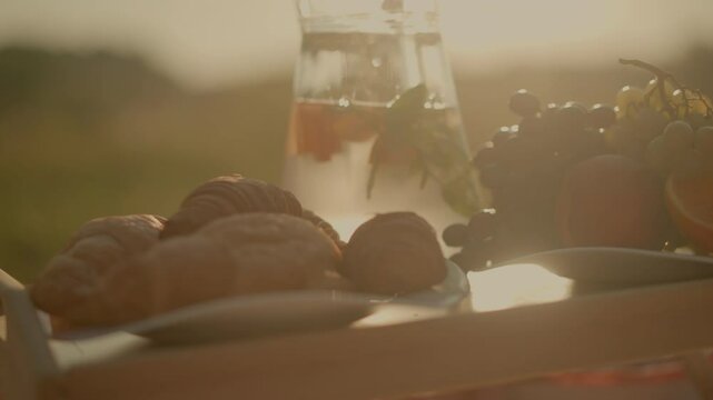 Close-up of rustic picnic setup on wooden table featuring fresh croissants, green grapes, and assorted fruits like orange slices and peach, alongside water pitcher under golden