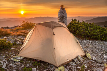 Young woman wrapped in a sleeping bag watches sunrise outside camping tent. Weekend adventure, people enjoy nature Carpathian Mountains.