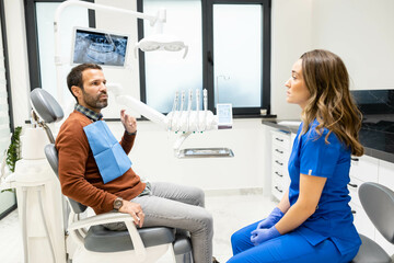 A dentist in blue scrubs listens to a seated patient in a clinic, surrounded by dental instruments and screens, representing a professional healthcare environment.