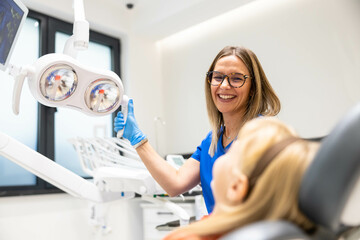 A dentist wearing glasses and gloves interacts with her young patient in a modern and brightly lit...