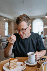 Man with glasses savors a bite of layered cake with a candied orange slice, accompanied by a cup of coffee in a warm and inviting cafe setting