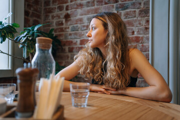 Blonde woman with curly hair waits at a cozy restaurant table, looking out the window in a peaceful urban cafe setting. She appears content and cheerful, soaking in the serene ambiance