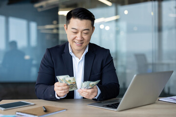 A young Asian man in a business suit is sitting in the office at the desk and counts the bills of...