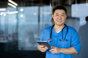 Portrait of a young Asian male doctor and medical staff standing in a clinic in a uniform with a stethoscope, holding a tablet and smiling at the camera