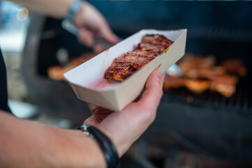 A close-up of hands holding a paper tray with grilled ribs, with a barbecue grill and another...