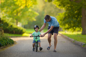 Young adult father teaching happy beautiful little boy to ride on first bike without pedals on sidewalk at city park. Learning to keep balance. Warm summer day. Cute 2 years old toddler. Front view.