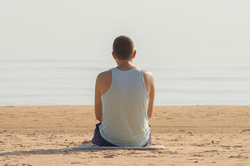 Young adult man sitting alone on sand and looking far away at sea beach in warm sunny summer day. Thinking about life. Back view.