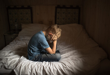 Sad unhappy little boy at home sitting on bed with head own
