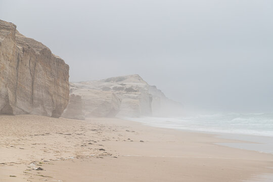 Atmospheric view of rocks and ocean with huge waves. Foggy Ocean Beach with Sand and Rocky Cliffs. 