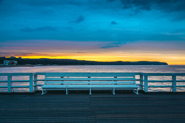 Fototapeta premium The inviting white bench stands on sopot pier at sunrise, offering a peaceful view of the colorful sky and tranquil baltic sea, creating a serene moment in the popular polish resort town