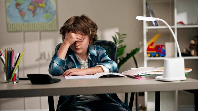 in the children's room a schoolboy in a white sweater and a blue checkered shirt at a desk doing homework on his studies writing difficult task children's suffering