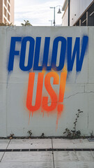 'FOLLOW US!' graffiti sprayed onto a white wall with bold blue and orange colors, set in an urban street setting with plants growing at the wall's base.

