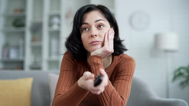 Lonely sad young adult woman bored switching television channels with remote control sitting on sofa in living room at home. Tired female has nothing interesting to watch on TV. Close up. Front view