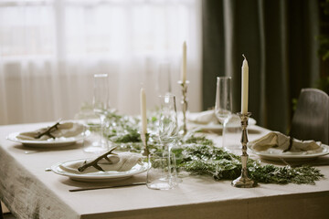 Medium close up of holiday table decorated with pine garland and polished silverware illuminated with sunrays through window in dining room