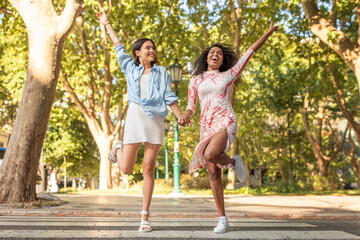 Fototapeta premium Two young women leap joyfully, holding hands in a vibrant park filled with lush trees. Their cheerful expressions and playful attire reflect a spirit of friendship and freedom.