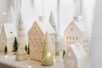Merry Christmas! Stylish Christmas miniature holiday village with white houses, trees and golden lights bokeh on snowy window. Atmospheric magical scene. Christmas holiday banner.
