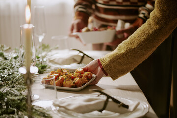 Unrecognizable female hand serving table with plate full of oven roasted potatoes while his husband helping her in blurred background