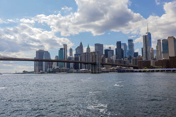 Fototapeta premium Brooklyn Bridge with Manhattan skyline and Statue of Liberty on Hudson River, under partly cloudy sky.